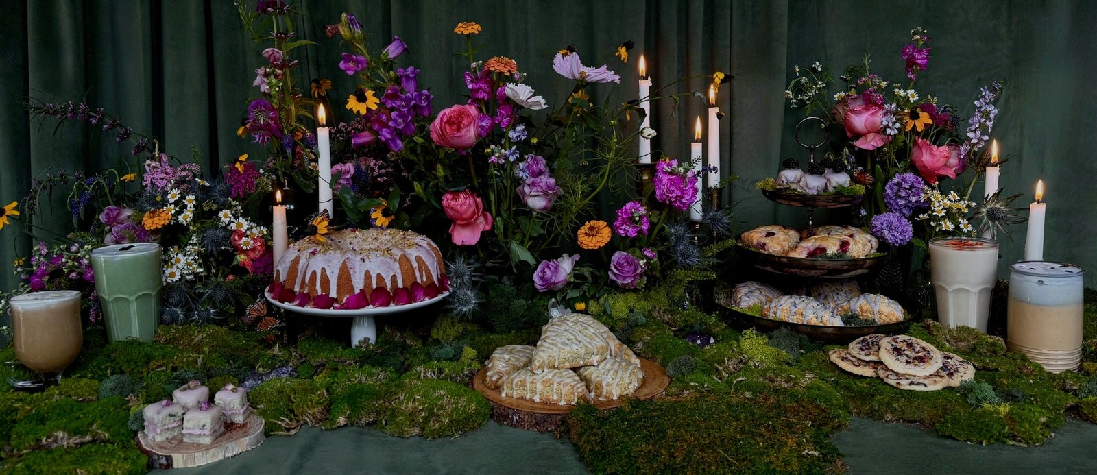 A Witches Brew catering spread: house-baked bundt cake, glazed scones, tea cakes, and cookies among wildflowers and candles, with matcha and lattes