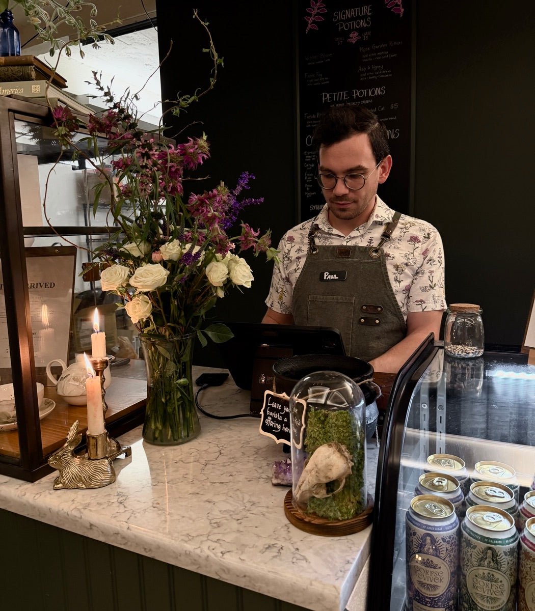 Paul DeSana behind the register at Witches Brew Coffee Co., beside fresh flowers and the chalkboard menu