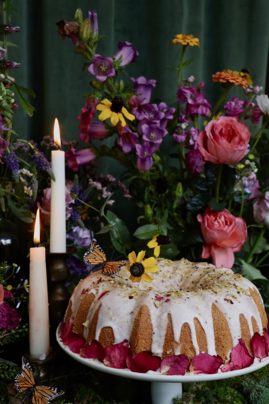 A glazed bundt cake ringed with rose petals among candles and wildflowers